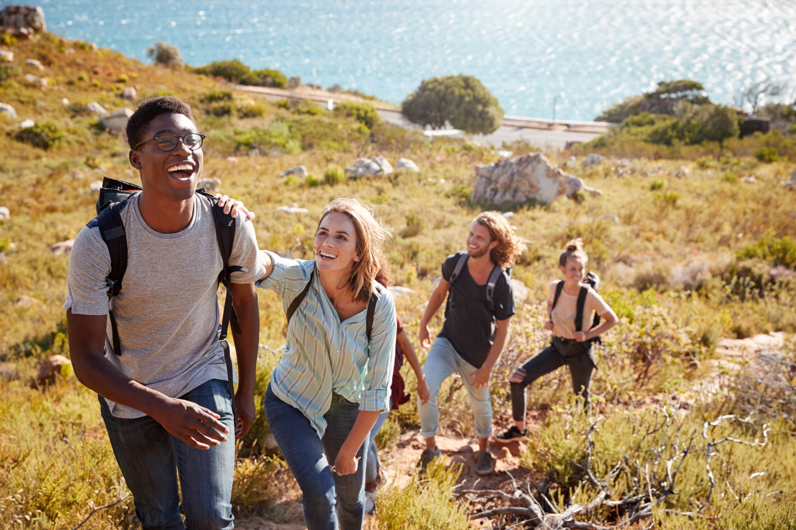 Millennial African American man leading friends hiking single file uphill on a path by the coast