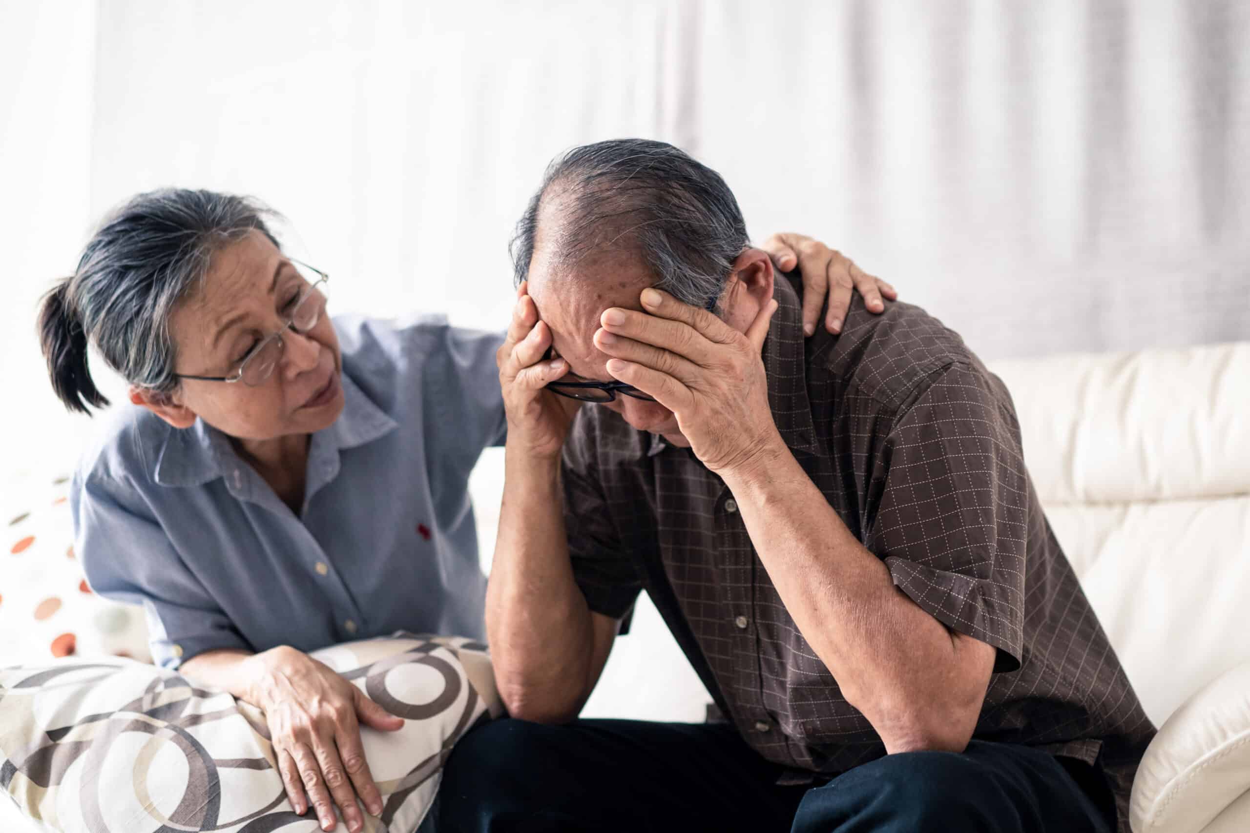 Asian senior old man having headache stress problem put hand on temple sitting on sofa at home. Elder wife sit beside consoling husband put hug, put hand on shoulder. Make comfort talking to relax him