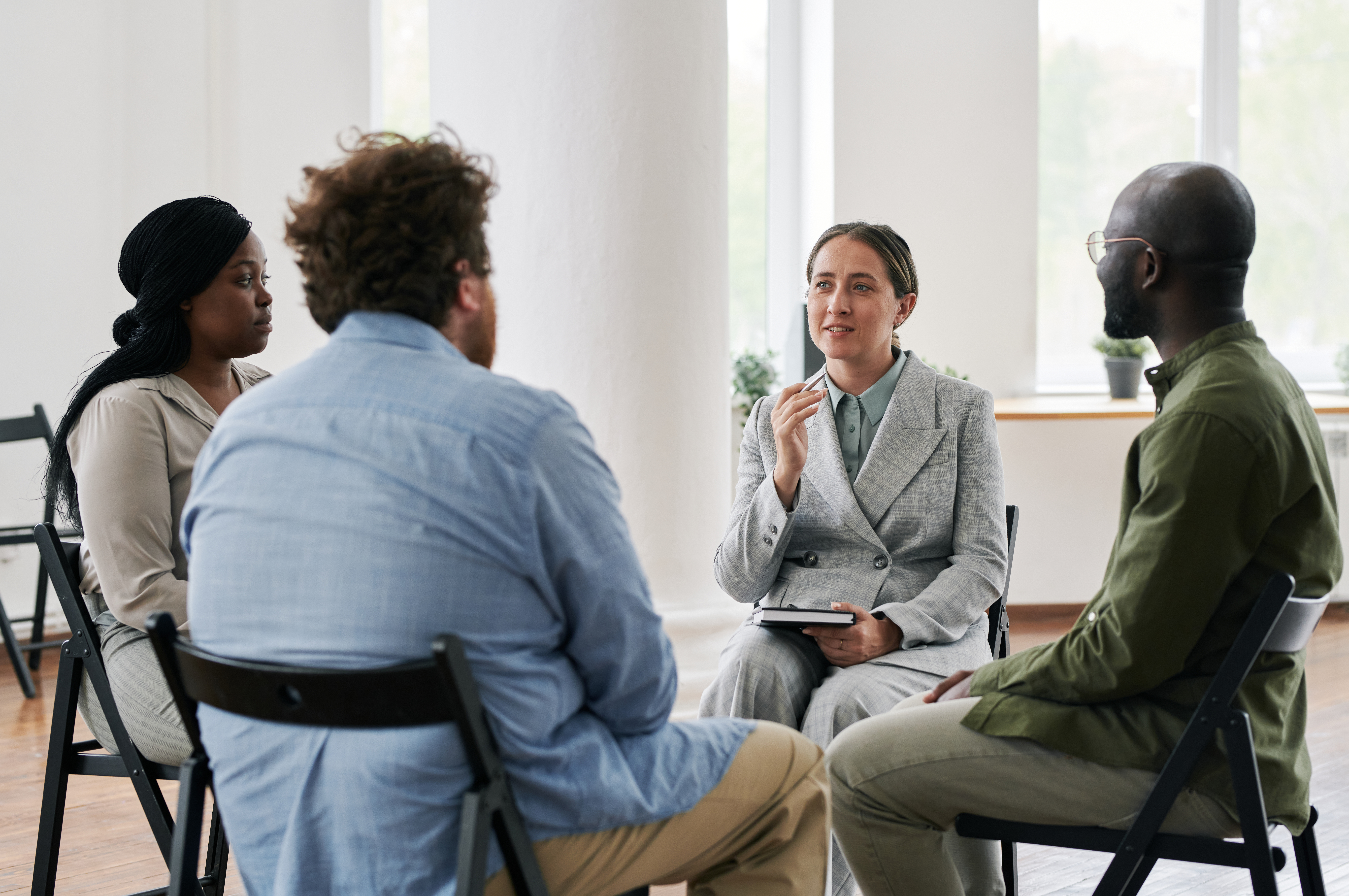 Young confident female psychologist in suit talking to group of intercultural people with psychological problems at session