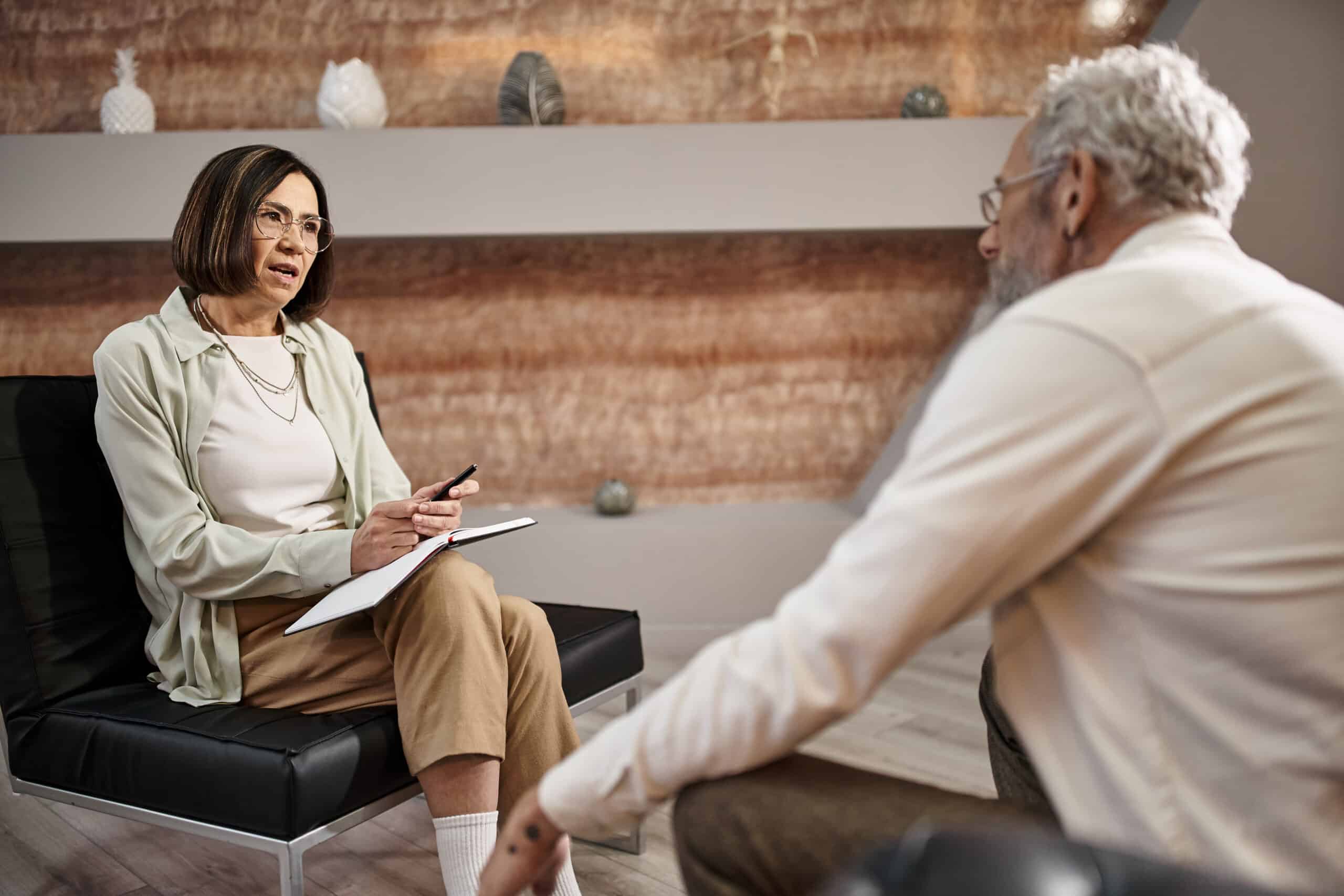 middle aged psychologist in glasses sitting and talking to bearded man during therapy session
