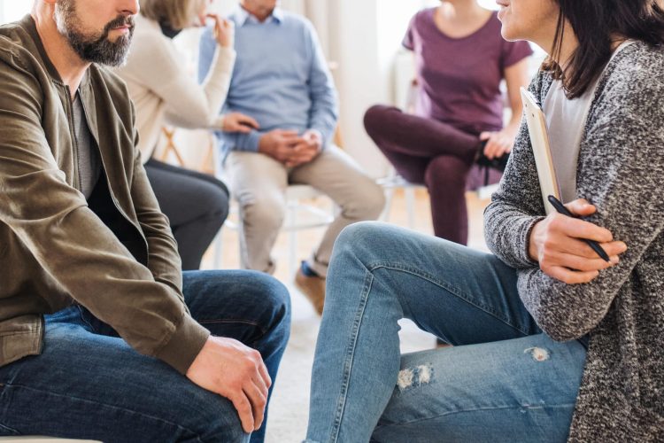 man in group therapy in a circle talking to woman therapist holding a clipboard